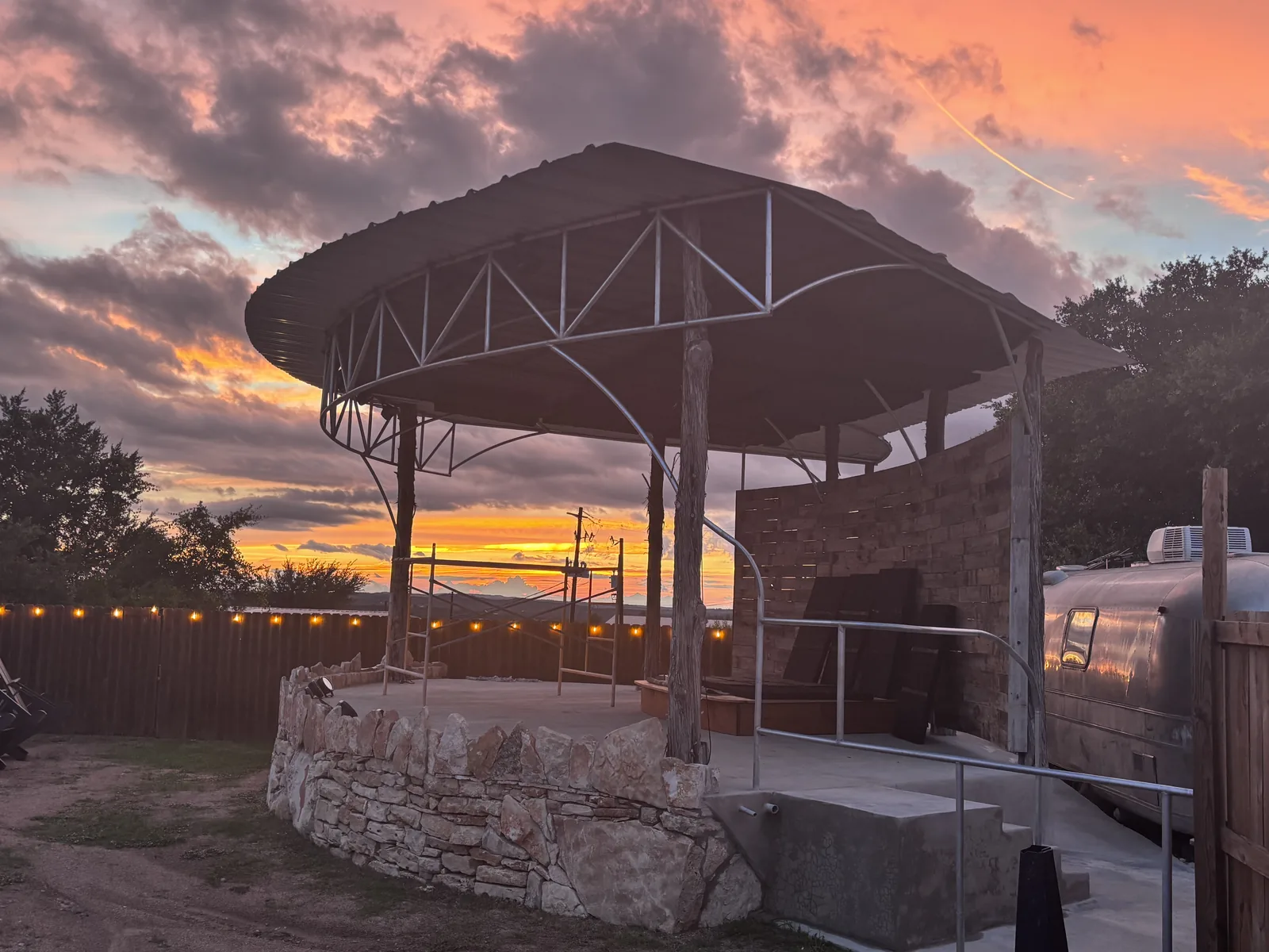 Sunset over the outdoor stage at Poodie's, with string lights along the fence and a pink and orange Hill Country sky behind the hills